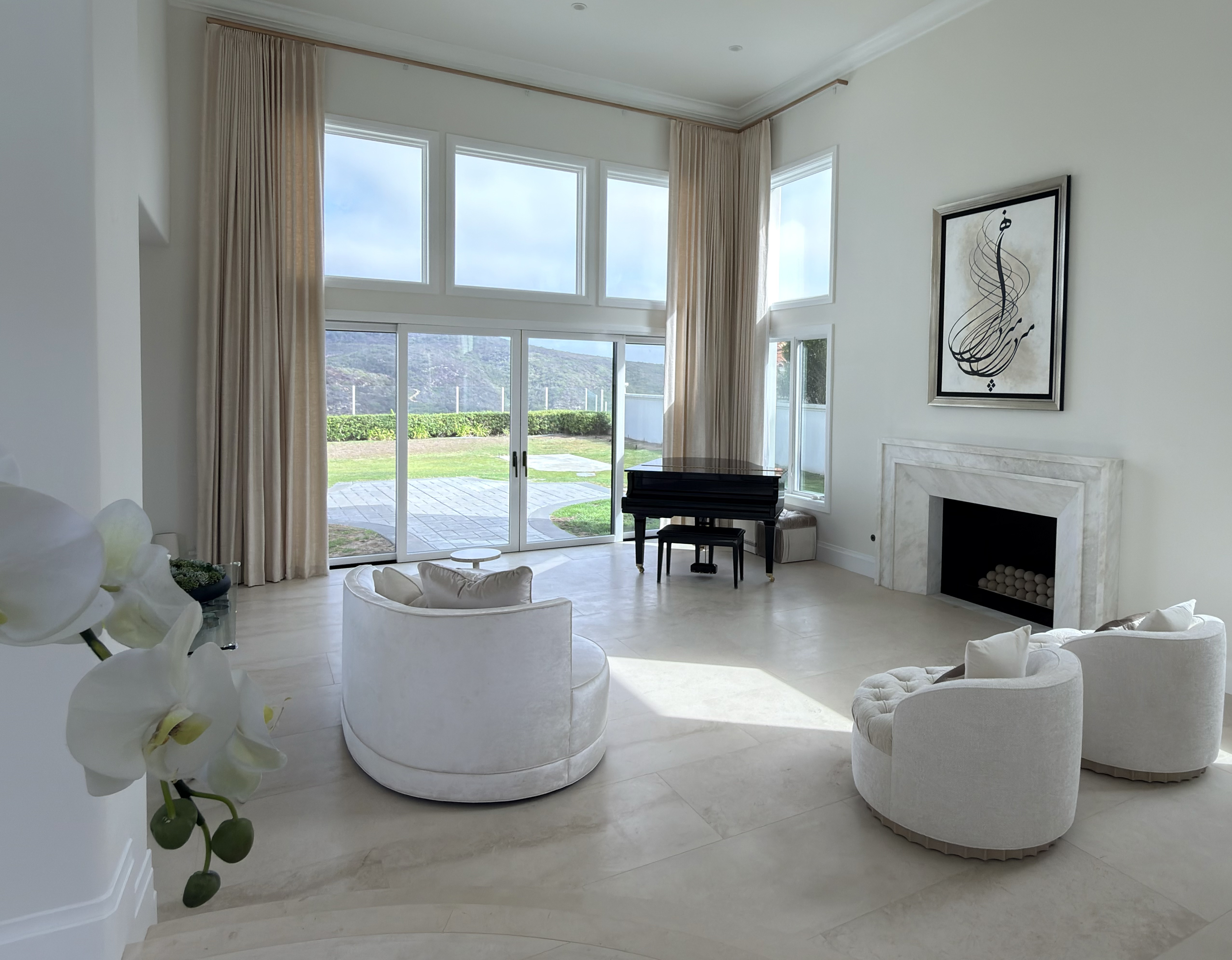 Light-filled living room with white lounge chairs, a grand piano, and a marble fireplace